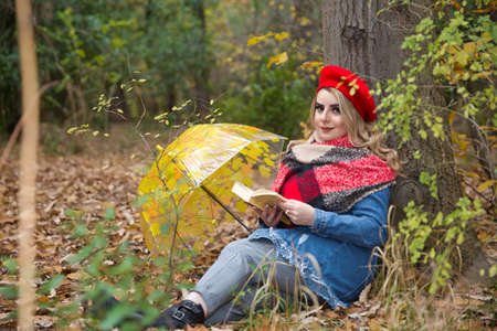Baku Azerbaijan 21.12.2019 .In the fall, the girl sits on the floor in the park and reads a book, with an umbrella next to it. The girl in the red hat . The girl smiles and looks at the cameraのeditorial素材