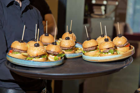 Catering service. Waiter carrying a tray of appetizers. Outdoor party with finger olives, burgers, sliders. man hold tray with burger at restaurant. Hands hold a tray with a cheeseburger.の写真素材