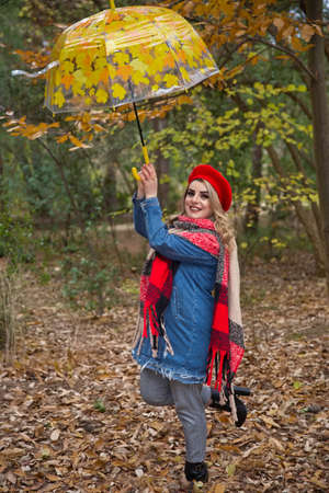 Baku Azerbaijan 21.12.2019 . Park between the leaves in front of the tree in autumn. The girl in the red hat is holding an umbrella, smiling, raising one of her legs and looking at the camera.のeditorial素材