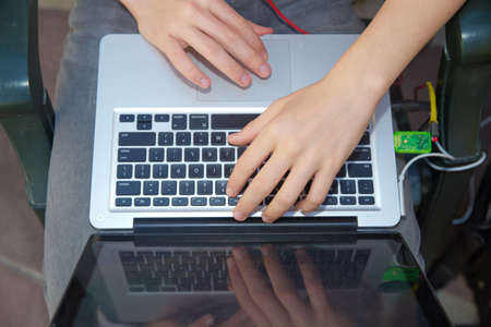 The boy is working on a computer on his lap . A cute Caucasian boy sitting on the bench in the park with his laptop on his knees and laughingの写真素材