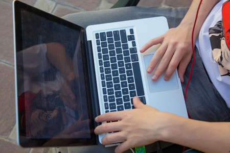 The boy is working on a computer on his lap . A cute Caucasian boy sitting on the bench in the park with his laptop on his knees and laughingの写真素材