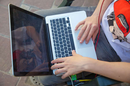 The boy is working on a computer on his lap . A cute Caucasian boy sitting on the bench in the park with his laptop on his knees and laughingの写真素材