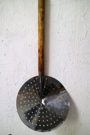 A vintage metal skimmer with a wooden handle, hanging against a textured white wall. The skimmer features a round, perforated design, ideal for draining liquids from food.の写真素材