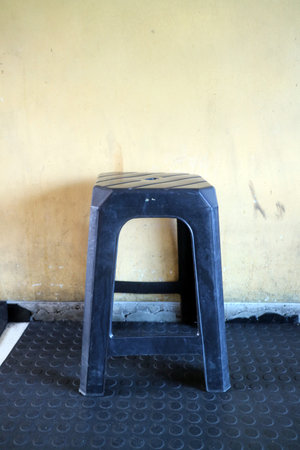 A simple black plastic stool against a yellow wall. The stool has a textured top and is positioned...