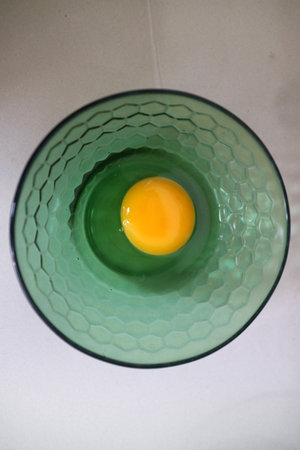 A raw egg yolk in a green glass bowl on a white surface.の写真素材