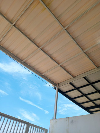 Upward view of a metal roof canopy with beams over a terrace, blue sky and clouds in background,...