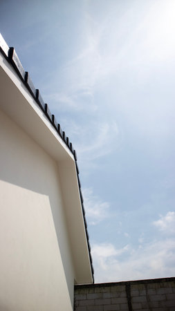 Low angle view of a modern white building roof edge against bright blue sky with sun flare and wispy...