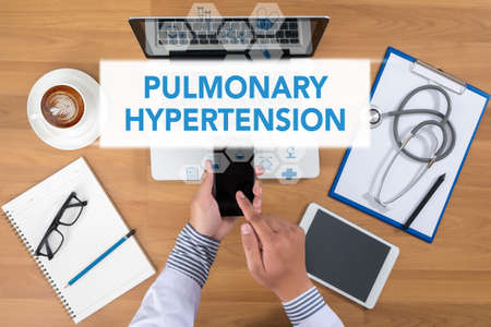 PULMONARY HYPERTENSION Doctor working at office desk and using a mobile touch screen phone, computer and medical equipment all around, top view, coffeeの写真素材