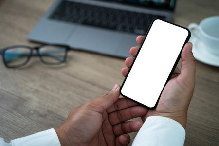 A person is holding a cell phone with a white screen. The phone is on a desk with a cupの写真素材
