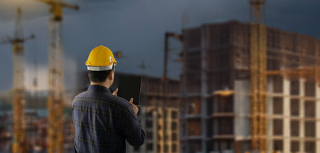 engineer working at site of a large building man wearing a yellow hard hat is looking at a tablet...