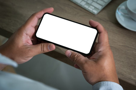 A person is using a laptop and smartphone at a modern office desk, holding a phone in one hand and typing on a keyboard, displaying digital communication and business with a white screenの写真素材
