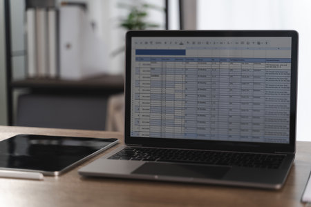 A report file laptop displaying spreadsheet with data and charts wooden desk modern office environment, with tablet and pen nearby, creatingの写真素材