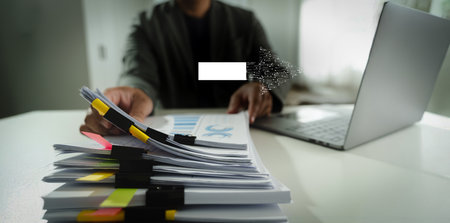 A digital transformation person working desk with laptop and large stack of documents, holding pen, modern office environment, with focus paperworkの写真素材