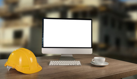 A laptop is open on a desk with a yellow hard hat and a white cup. The laptop is empty and the desk is cluttered with construction tools.の写真素材