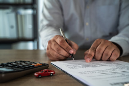 An insurance on paperwork assurance person gray shirt is signing document wooden table, with calculator and small red car model nearby, suggestingの写真素材