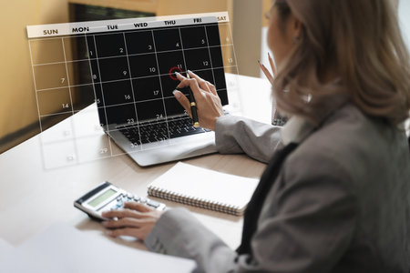 A 2025 calendar woman business suit is planning her schedule digital calendar overlay, using pen and laptop, with calculator and notebook nearbyの写真素材