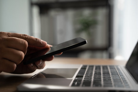 Hand operating a smartphone beside a laptop in an indoor workspace, using smartphone remote work and...
