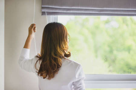 Rear view of a young woman wearing a robe and holding the curtains open to look out of a large light window at home, interior. Positive and aspirational lifestyle. Woman looking out a window, indoors.の写真素材
