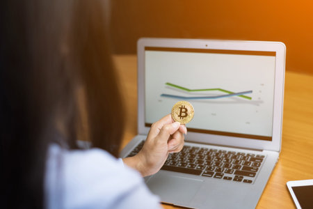 Business woman's hand holding a gold bitcoin at office with sun effect.Background graph on laptop.