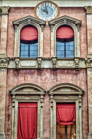 Traveling. Italian renaissance architecture. Building facade adorned with a clock. Embellished windows with hanging red drapes. Bologna, Italy.の写真素材