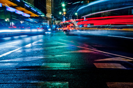 A bustling city street at night, captured in a long exposure. Vehicle lights streak across the wet asphalt, illuminating pedestrian crossings. Urban energy in motion.の写真素材