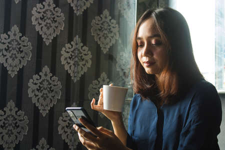 Relaxed Smilling Asian Woman drinking coffee while using a cell phone for workの写真素材