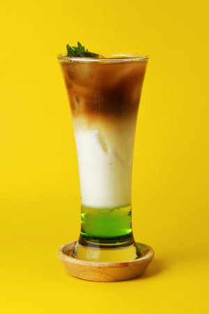 Closeup photo of a pandan iced coffee drink layered with melon syrup, milk and chocolate coffee in a tall glass on a yellow backgroundの写真素材