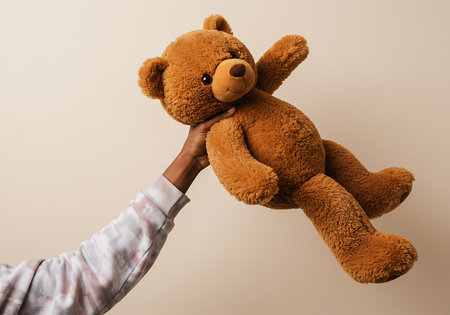 A close-up shot of a hand holding a large, brown teddy bear against a light beige background. Generative Aiの素材