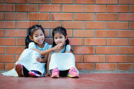 Asia little girl sitting with friend on brick wall backgroundの写真素材