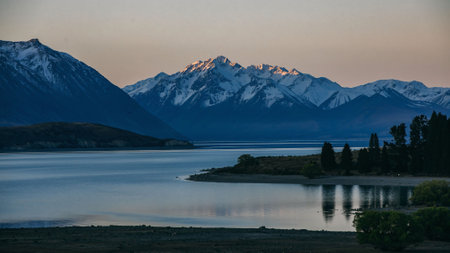 As dusk sets in, the last rays of sunlight accentuate the snowy peaks towering over a calm lake, with the silhouettes of pine trees adding to the tranquil scene.の写真素材