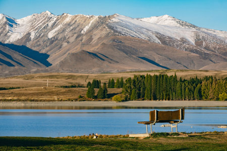 A solitary bench overlooking a calm lake offers a perfect spot for contemplation, with the impressive backdrop of snow-dusted mountains and lush pine trees.の写真素材