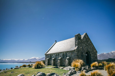 A quaint stone chapel with a slate roof stands serenely by a lake, set against a backdrop of distant snow-capped mountains and a clear blue sky.の写真素材