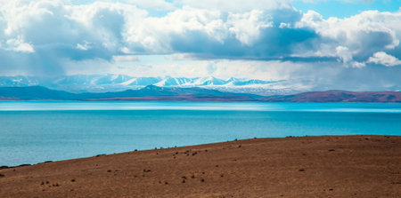A breathtaking view of snow-capped mountains beneath a dramatic sky, reflecting in the tranquil blue waters of a vast lake, bordered by a rugged brown shoreline.の写真素材