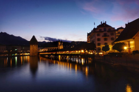 A stunning evening view of the Chapel Bridge in Lucerne, Switzerland, with its illuminated wooden structure reflecting on the calm water of the Reuss River.の写真素材