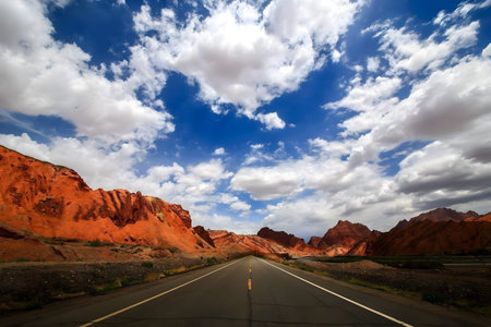 A straight road cuts through a dramatic red rock canyon under a vibrant blue sky filled with...