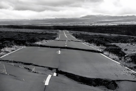A black and white photograph depicting a cracked and broken road stretching through a barren landscape. The road appears to be abandoned, surrounded by rocky terrain and under a cloudy sky, evoking a sense of desolation and isolation.の写真素材