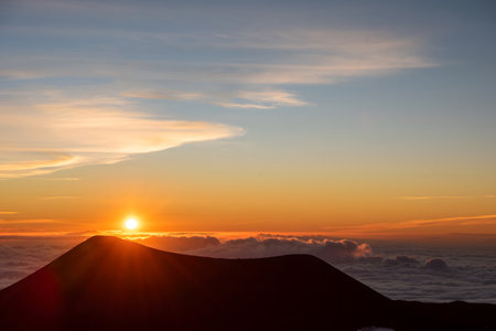 A breathtaking view of the sun rising above the horizon, casting warm hues across the sky and illuminating the clouds below. The silhouette of a mountain peak is prominently featured in the foreground, creating a stunning contrast against the vibrant colors of dawn.の写真素材
