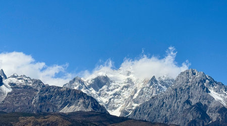 A breathtaking view of towering snow-capped mountains under a clear blue sky, with wispy clouds gently resting on the peaks. The rugged terrain showcases a mix of rocky surfaces and patches of greenery, highlighting the natural beauty of the mountainous landscape.の写真素材