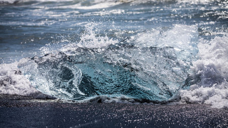 A block of clear ice with air pockets is washed by waves on the black sands of Diamond Beach on...