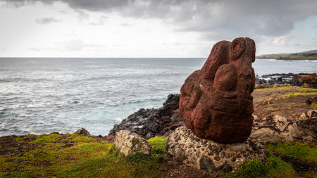 An old, historic red sculpture, made of red volcanic rock, stands on a beige pedestal on the coast of Easter Island above the Pacific Ocean.の写真素材