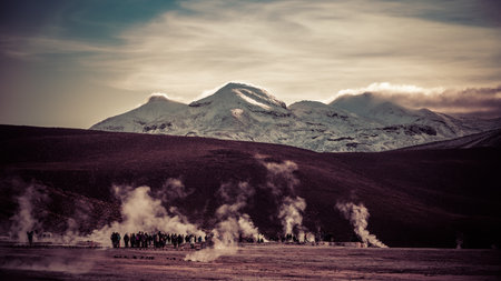 Snow-covered peak in the golden light of sunrise and a crowd of people amid clouds of steam from hot springs.の写真素材