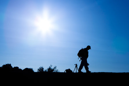 A silhouette of a professional photographer setting off on an adventure with a camera, tripod and backpack. Sun and blue sky in the background.の写真素材