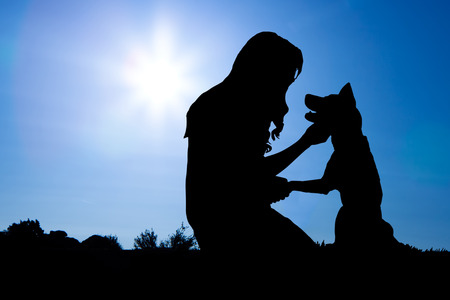 Silhouette of a woman with long hair sitting with a dog and shaking his handの写真素材