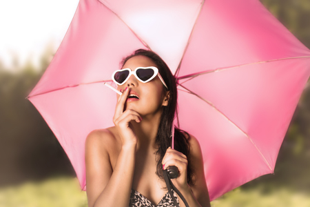 Mexican black haired woman smokes in a garden wearing a bikini and protecting herself from the sun with an umbrellaの写真素材