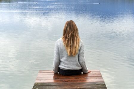 Young blond girl with long hair, sit down and back to the camera on a wooden pier in front of a calm lake looking to the horizon in Baviera. Travel concept.の写真素材