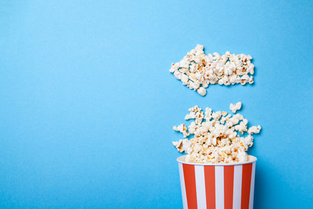 Spilled popcorn in the form of an arrow to the left and paper bucket in red strip on blue background. Copy space for textの写真素材