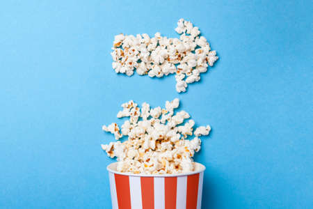 Spilled popcorn in the form of an arrow to the right and  paper bucket in red strip on blue background.の写真素材