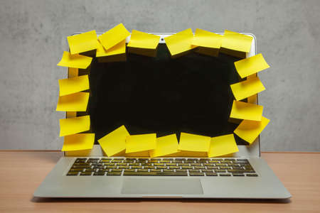Laptop in reminders with yellow stickers notes on the display frame on table with gray wall. Many unfulfilled tasks, deadlineの写真素材