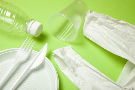Plastic disposable utensils on green background. fork, knives, plates, cups and a package, bottle, ...