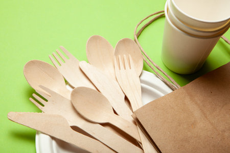 Eco-friendly disposable utensils made of bamboo wood and paper on a green background. Draped spoons,...